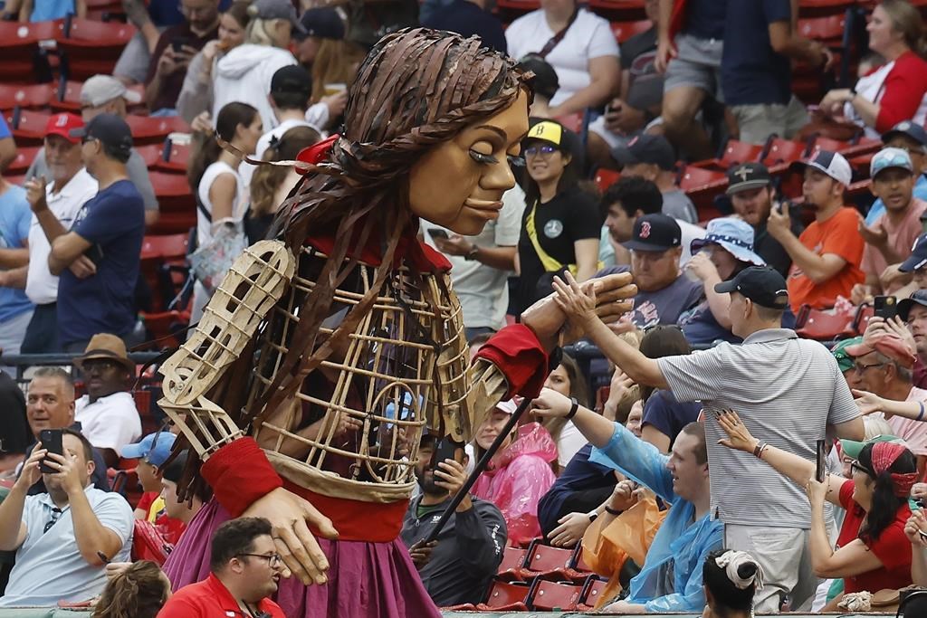 Little Amal, , left,a 12-foot puppet of a Syrian refugee, interacts with fans at Fenway Park before a baseball game between the Boston Red Sox and the Baltimore Orioles, Saturday, Sept. 9, 2023, in Boston. The puppet is visiting key historic places in the United States to raise awareness about immigration and migration. (AP Photo/Michael Dwyer)